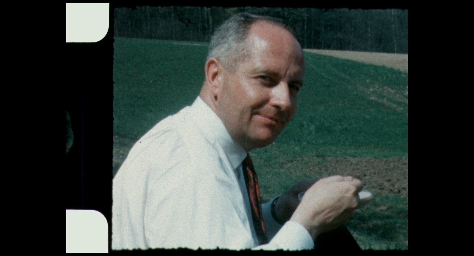 Körnige Filmaufnahme von einem Mann im Halbprofil, der in die Kamera lächelt. Er trägt ein helles Hemd und eine Krawatte.