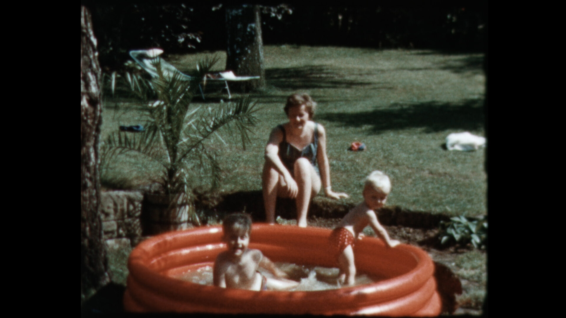 Körnige Aufnahme aus einem Garten. Auf dem kurz gemähten Rasen steht ein rotes, aufblasbares Planschbecken, in dem zwei Kinder im Kindergartenalter planschen. Hinter dem Becken sitzt eine Frau im Badeanzug und beobachtet die spielenden Kinder.
