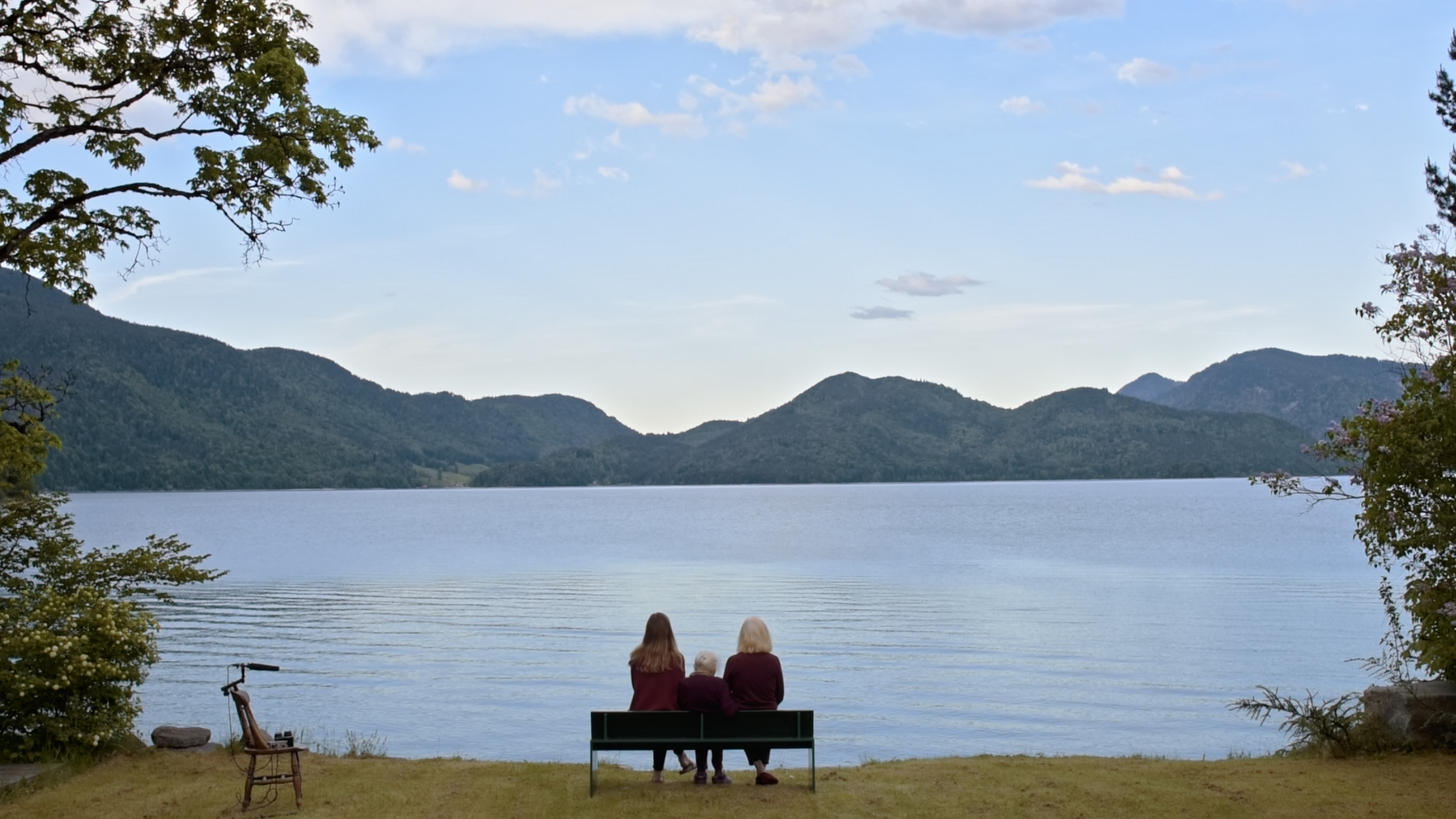 Panoramablick auf See vor Gebirge, auf einer Bank am Ufer sitzen drei Frauen unterschiedlicher Generationen.